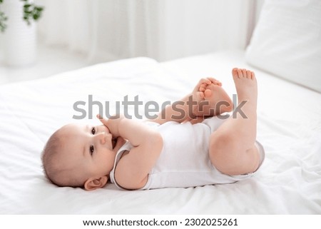a cute little boy in a white bodysuit is lying on his back on a bed on white bedding in the bedroom, playing with his arms and legs, portrait of a cute child with brown eyes and blond hair Royalty-Free Stock Photo #2302025261