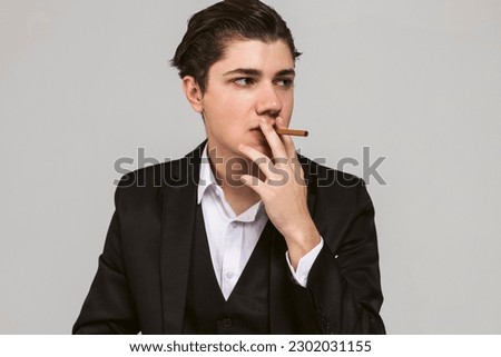 Portrait of a young mafia member with a slicked back hair, dressed in black suit smoking a cigar, throws a menacing gaze. Italian mafia gangster style. isolated on white background. Royalty-Free Stock Photo #2302031155