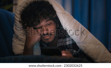 Sad sleepy guy Latino tired man upset Arabian sleeping napping boring Indian male at night evening dark home under blanket cover with duvet scrolling mobile phone addict smartphone bored social media Royalty-Free Stock Photo #2302032625