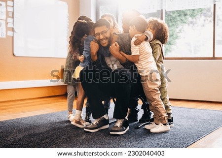 Teacher and students greet each other excitedly. Elementary school educator smiling at the camera as his class embraces him. Group of children celebrate reuniting with their tutor in a primary school. Royalty-Free Stock Photo #2302040403