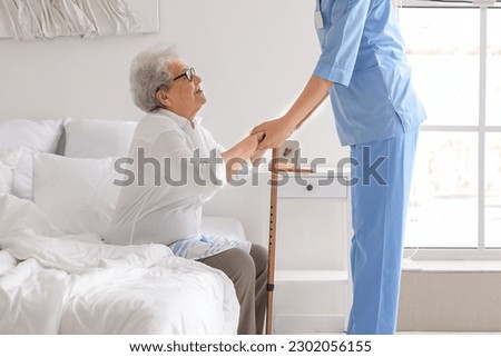 Young caregiver helping senior woman with stick to stand up in bedroom Royalty-Free Stock Photo #2302056155