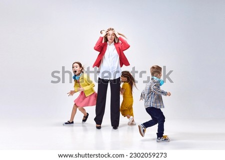 Portrait of tired young woman, mother holding head with hands around little noisy kids playing against grey studio background. Concept of family, motherhood, childhood, fashion, lifestyle
