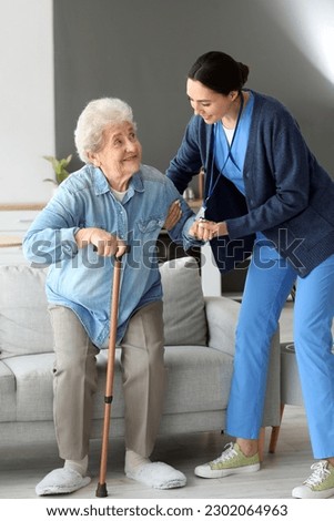 Young caregiver helping senior woman with stick to stand up at home