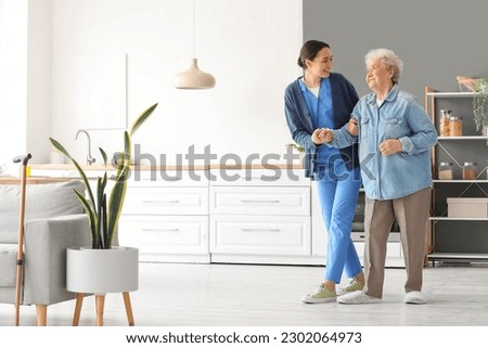 Young caregiver helping senior woman to walk in kitchen