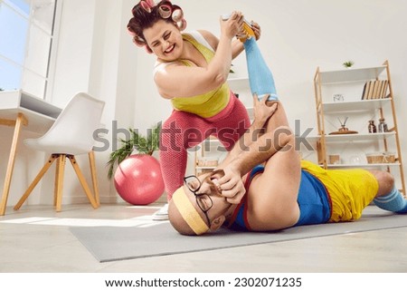 Funny fat young woman makes man do leg stretching exercise. Chubby wife makes husband do painful stretch. Happy overweight woman tortures her sports trainer as act of vengeance for difficult workouts