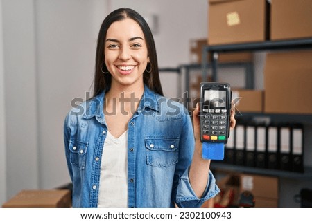 Hispanic woman working at small business ecommerce holding credit card and dataphone looking positive and happy standing and smiling with a confident smile showing teeth 