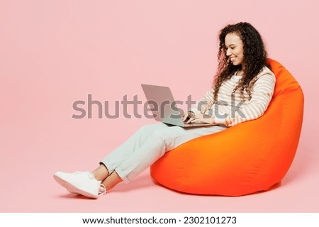 Full body fun young IT woman of African American ethnicity she wear light casual clothes sit in bag chair hold use work on laptop pc computer isolated on plain pastel pink background studio portrait