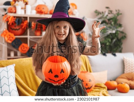 Adorable blonde girl wearing witch costume doing fear gesture with hands at home