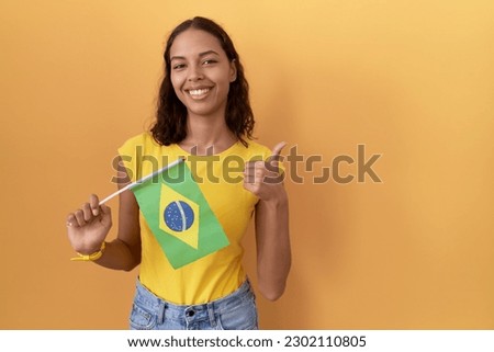 Young hispanic woman holding brazil flag doing happy thumbs up gesture with hand. approving expression looking at the camera showing success. 