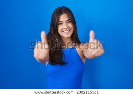Hispanic woman standing over blue background approving doing positive gesture with hand, thumbs up smiling and happy for success. winner gesture. 
