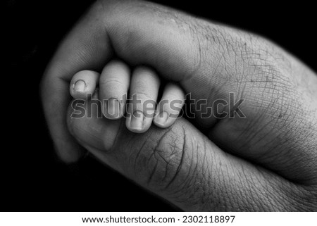 Close-up of a small hand of a child and the hand of mother and father. A newborn baby after birth holds tightly, squeezes the thumb of its parents. Black and white photography on a black background.