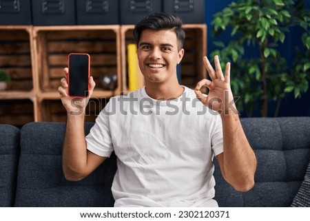 Hispanic man holding smartphone showing blank screen doing ok sign with fingers, smiling friendly gesturing excellent symbol 