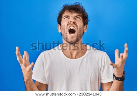 Hispanic young man standing over blue background crazy and mad shouting and yelling with aggressive expression and arms raised. frustration concept.  Royalty-Free Stock Photo #2302141043