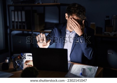 Hispanic young man working at the office at night covering eyes with hands and doing stop gesture with sad and fear expression. embarrassed and negative concept. 