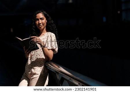 Woman leaning on banister holding a book and looking away. She is wearing a spring blouse and trousers and a lot of jewelry on her hands. Curly hair.