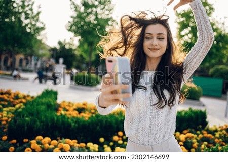 A chic young boho brunette is standing on a city street downtown and taking selfies while smiling at the phone on a sunny summer day. A young fashionable woman is taking self-portraits on the street.