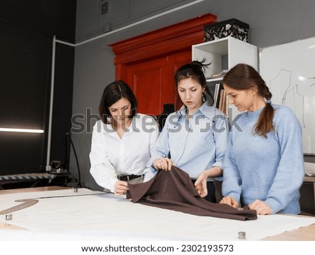Three female fashion designers work together in a tailoring studio. A group of women dressmakers discusses fabric, pattern and sketch in a sewing workshop. The concept of small business