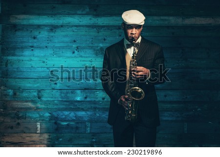Vintage african american jazz musician with saxophone in front of old wooden wall. Wearing suit and cap. Royalty-Free Stock Photo #230219896