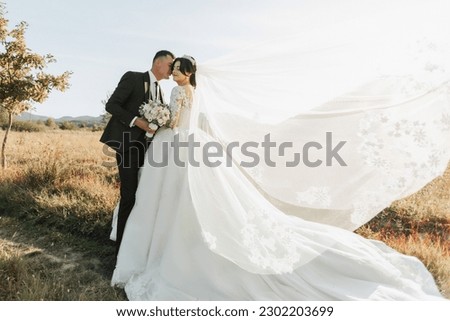 Wedding photo. A groom in a black suit and a bride in a white dress and veil blown by the wind stand kissing in a field against a background of trees and large mountains. Photo in a light key. Royalty-Free Stock Photo #2302203699