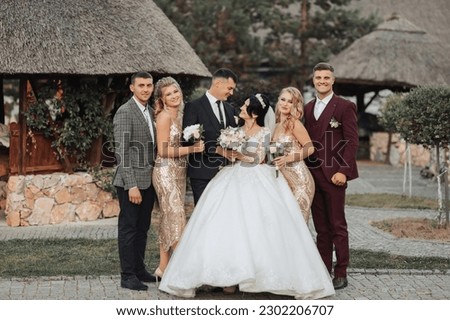  Front view of newlyweds standing and hugging, between friends. Girls in golden dresses hold bouquets, the bride in a voluminous dress, the groom and his friends in suits. Wedding in nature