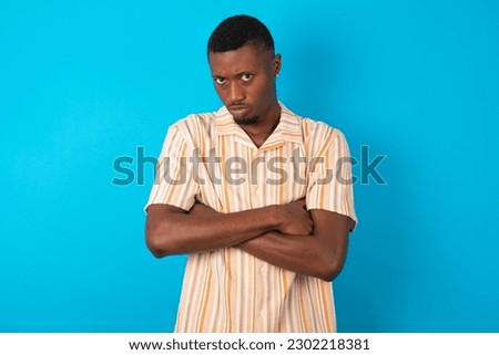 Gloomy dissatisfied young man wearing Hawaiian striped shirt over blue background looks with miserable expression at camera from under forehead, makes unhappy grimace