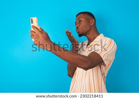 young man wearing Hawaiian striped shirt over blue background blows air kiss at camera of smartphone and takes selfie, sends mwah via online call.