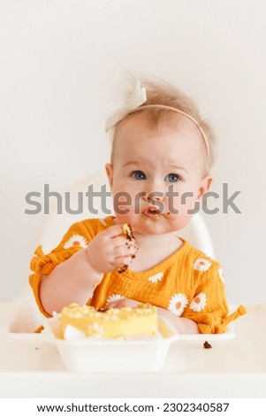 A charming little one-year-old baby girl in an elegant dress with daisies is sitting on a high chair and eating a cake. Celebrate one year of birth.