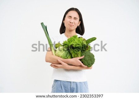 Miserable asian woman, holds green vegetables and complains at het diet, looks at calories, white background.