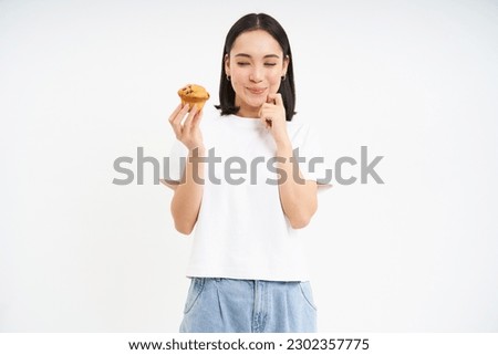 Image of young modern woman with cupcake, looks at delicious pastry from bakery with happy face, white background.