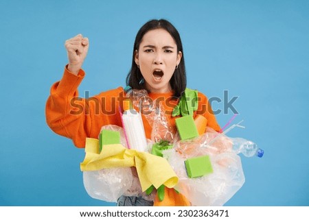Angry Korean woman shouts, holds plastic waste, urges to recycle garbage, shakes fist, blue background.