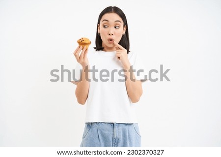 Image of young modern woman with cupcake, looks at delicious pastry from bakery with happy face, white background.