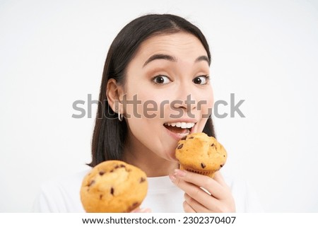 Image of happy korean girl who loves pastry, smiling and eating cupcakes, isolated on white background.