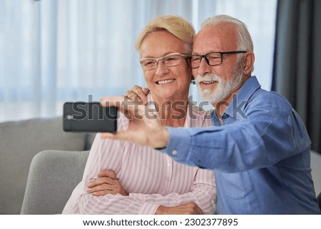 Happy elderly couple taking selfie, having fun with phone camera or recording video for vlog. Smiling aged wife and husband hugging, looking at camera, posing for photo
