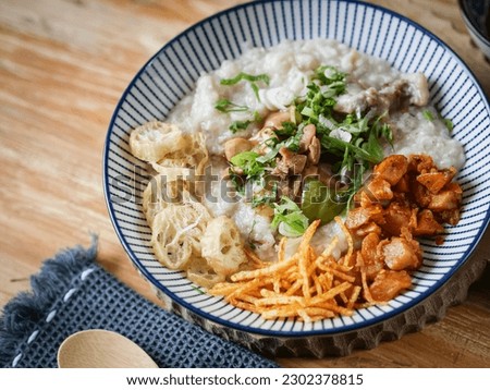Bubur ayam or chicken porridge is Chinese Indonesian rice porridge topped with various savoury condiments served on white bowl and wooden table. Royalty-Free Stock Photo #2302378815