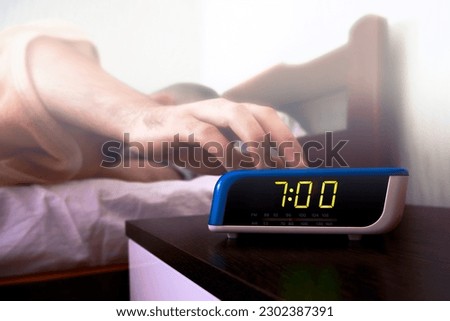 A sleeping man's hand reaches for a digital alarm clock on the nightstand. Color toning and haze filter on the image