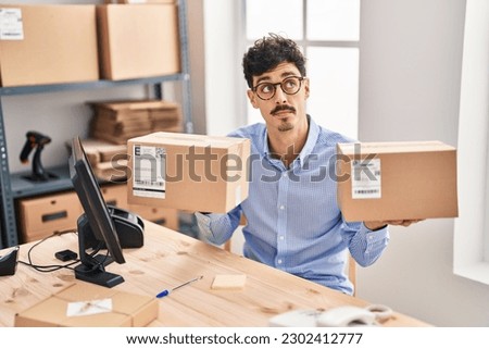 Hispanic man working at small business ecommerce holding packages smiling looking to the side and staring away thinking. 