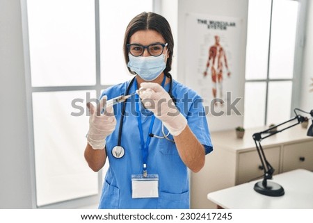 Young hispanic woman wearing doctor uniform holding covid-19 vaccine at clinic