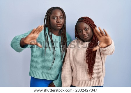 Two african woman standing over blue background doing frame using hands palms and fingers, camera perspective 