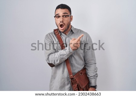 Young hispanic man wearing suitcase surprised pointing with finger to the side, open mouth amazed expression. 