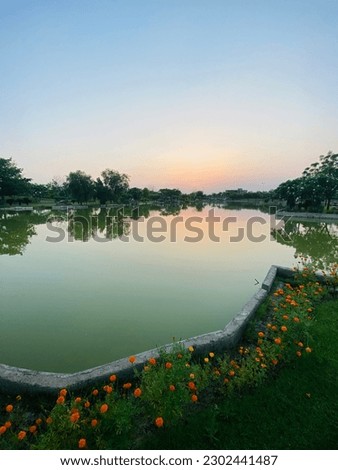 Picture shows lake with sunset sky, green trees, grass, and orange flowers. Captures nature's beauty, tranquil and visually appealing. Striking contrast between sky and greenery, captivating image.