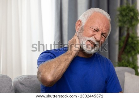 Elderly man suffering from neck pain sitting on sofa at home Royalty-Free Stock Photo #2302445371