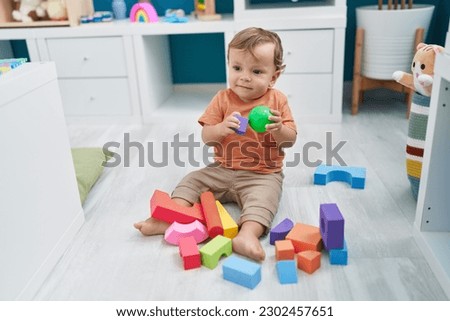 Adorable blond toddler playing with ball and geometry blocks sitting on floor at kindergarten