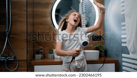 Beautiful blond little girl in bathrobe singing a song and dancing with a hair dryer. Child sings and dances in a shower after taking shower in bathroom. Royalty-Free Stock Photo #2302466195