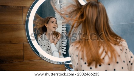 Beautiful blond woman in bathrobe singing a song and dancing with a hair dryer, looking at the mirror. Girl sings and dances in a shower after taking shower in bathroom. Royalty-Free Stock Photo #2302466233