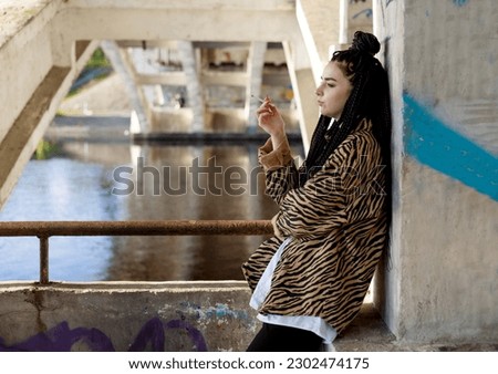 Thinker teenager in depression sitting down under the bridge at the sunset. Concept of teenage problems. Unhealthy habits. Royalty-Free Stock Photo #2302474175