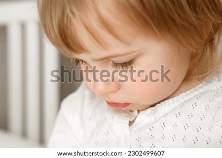 Portrait of two year old girl with a focused gaze. Small toddler child looks down unhappy. Blonde 2 years baby with white shirt looking with sad face. Upset little kid. Anger, aggression, resentment.