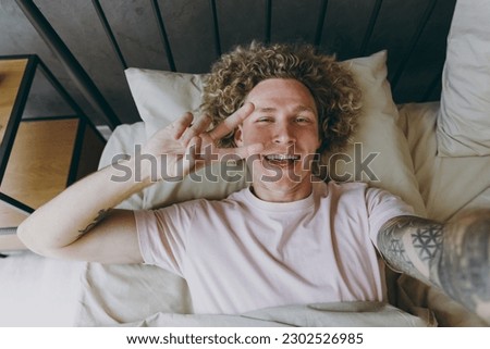 Close up young man wear white t-shirt he lying in bed do selfie shot pov mobile cell phone show v-sign rest spend time in bedroom home in own room wake up dream lost in good mood Real estate concept
