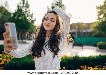 A cheerful young brunette is standing on a city street downtown and taking selfies while smiling at her phone. A boho woman is taking self-portraits with her phone while standing on the city square.