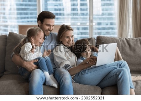 Happy couple of parents using online learning app on laptop, for educating little kids, resting on couch together, holding digital device, enjoying wireless technology, domestic internet connection