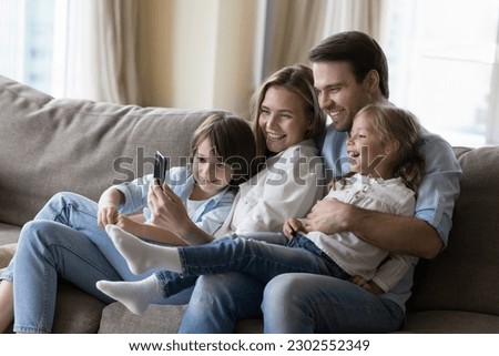 Young mother and father hugging cheerful kids on home sofa, taking happy family picture on smartphone, using mobile phone together, for online communication, enjoying domestic Internet technology